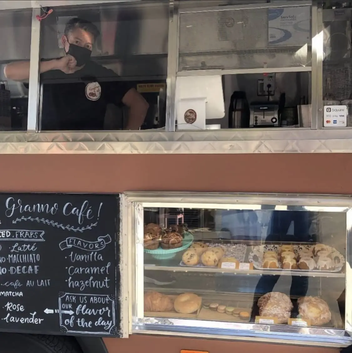 Paulo standing inside the Granno Cafe's ordering window. Below the window is a glass display of fresh pastries and a blackboard cafe menu.